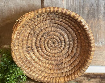 May include: A round, shallow basket constructed from coiled, natural-colored dried grasses and fibers. The weaving features a tight, circular pattern radiating from the center, with visible light and dark brown tones throughout the material.