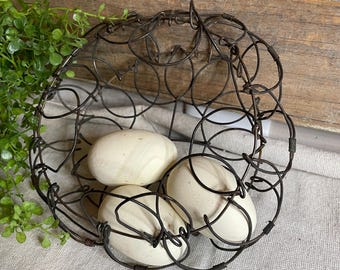 May include: A rustic, dark metal wire basket holds three cream-colored decorative eggs. The basket has a circular design with multiple wire loops and a handle. The eggs have a smooth, matte finish. The basket is placed on a neutral-toned fabric.