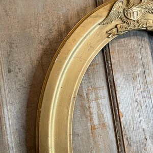 May include: Gold-colored oval picture frame with a decorative emblem featuring an eagle, shield, and the letters "U.S.A." The frame has a distressed finish, suggesting an antique or vintage style. The frame is made of wood.