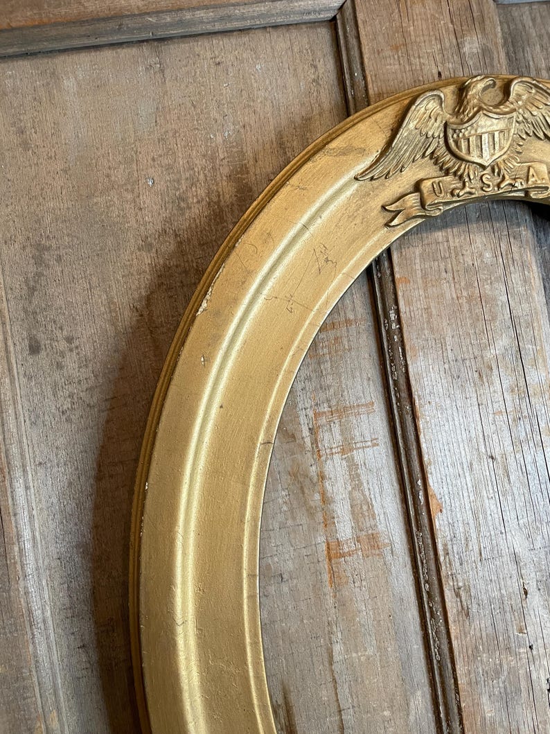May include: Gold-colored oval picture frame with a decorative emblem featuring an eagle, shield, and the letters "U.S.A." The frame has a distressed finish, suggesting an antique or vintage style. The frame is made of wood.