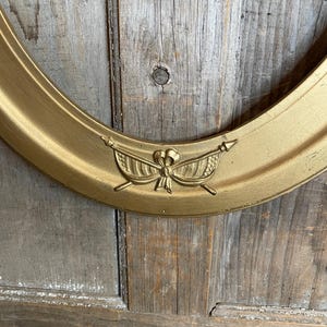 May include: Oval gold-colored picture frame with a decorative emblem at the top. The frame has a smooth, metallic finish and is set against a weathered wooden background. The emblem features a bow and arrows.