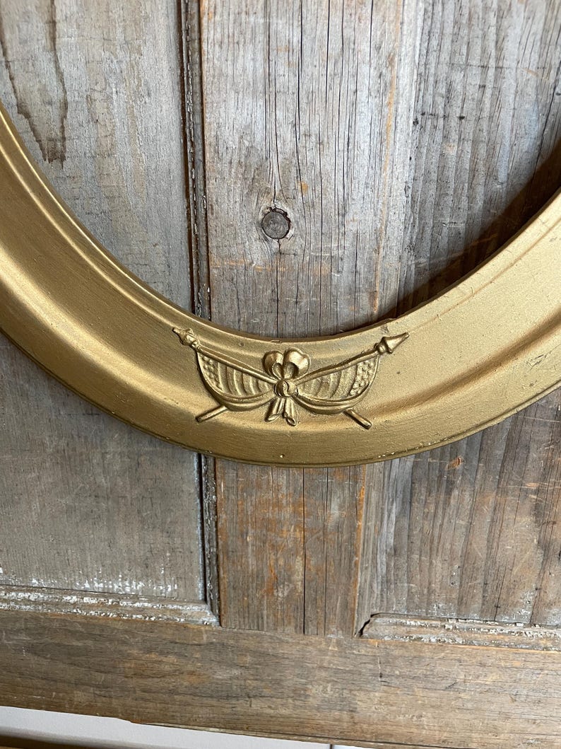 May include: Oval gold-colored picture frame with a decorative emblem at the top. The frame has a smooth, metallic finish and is set against a weathered wooden background. The emblem features a bow and arrows.