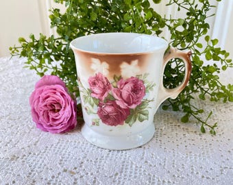 May include: A white ceramic mug featuring a brown-tinted upper band and a floral pattern of pink and deep red roses with green leaves. The mug has a curved handle and a flared base, accompanied by a single fresh pink rose placed beside it.