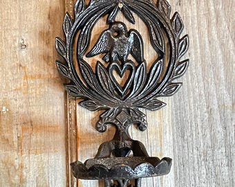 May include: A dark gray cast iron wall sconce with a bird and heart design. The sconce features a circular wreath, a small dish, and a decorative base. The background is a light brown wooden surface.