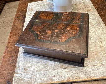 May include: A vintage wooden box with a hinged lid, featuring a dark brown finish with decorative accents. The box sits on a light-colored wooden surface. A white pitcher with greenery is in the background. The box appears to be antique.