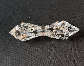 May include: A clear, faceted glass object with a double-pointed, symmetrical design. The piece features intricate, carved geometric patterns along its length, tapering to sharp points at both ends, with a central cylindrical section.