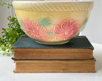 May include: A pale yellow ceramic mixing bowl featuring a raised, embossed floral pattern with pink petals and green leaves. The bowl has a thick, rope-like textured rim and a smooth, glossy finish, resting on a stack of two vintage hardcover books.