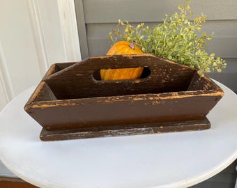 May include: A rustic, brown wooden tray with a handle, holding a small orange pumpkin and green foliage. The tray has a distressed finish, suggesting an antique or farmhouse style. The arrangement sits on a white table, evoking a fall or harvest theme.