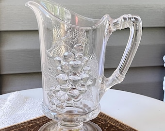 May include: Clear glass pitcher with a pedestal base and a handle. The pitcher features a raised grape design on the front. The pitcher is sitting on a vintage book.