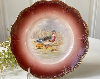 May include: A round ceramic plate with a scalloped edge featuring a deep red gradient border and a central illustration of a duck and a duckling standing in tall grass near water. The rim has a textured, embossed pattern with gold accents.