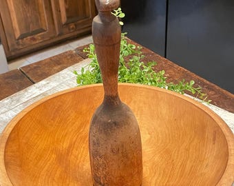 May include: A tall, cylindrical wooden pestle with a rounded top handle and a wide, flat base, positioned upright inside a large, smooth, circular wooden bowl with a natural light brown grain and a polished finish throughout the surface.