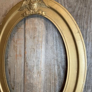 May include: Oval gold-colored picture frame with an embossed eagle and banner that reads "USA". The frame has a detailed design and is set against a rustic wooden background. A decorative piece for displaying photos or artwork.