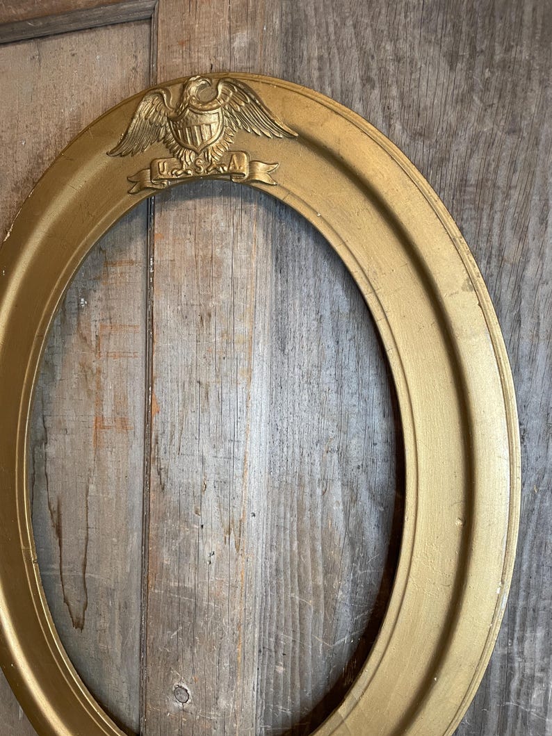 May include: Oval gold-colored picture frame with an embossed eagle and banner that reads "USA". The frame has a detailed design and is set against a rustic wooden background. A decorative piece for displaying photos or artwork.
