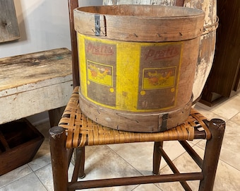 May include: An antique wooden chair with a woven seat supports a large, round wooden container. The container has a yellow label with the word "Pratts" and illustrations. The chair and container have a rustic, vintage aesthetic.