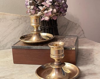 May include: Two polished brass candle holders with wide, circular drip trays and cylindrical sockets. The holders feature a smooth, reflective metallic finish and a tiered base design, positioned on a rectangular wooden box with a patterned lid.