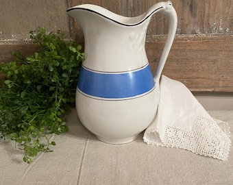 May include: White ceramic pitcher with a blue band and black trim. The pitcher has a curved handle and a spout. A white lace-edged cloth rests beside the pitcher. Green foliage is on the left side of the image.