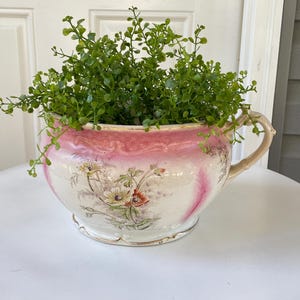 May include: A vintage, white and pink floral planter with a handle, filled with green foliage. The planter has a decorative floral design and a gold trim. The planter is sitting on a white table.