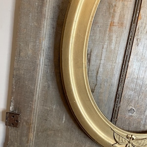 May include: A gold-colored, oval picture frame with a decorative emblem at the bottom. The frame is set against a weathered, wooden background. The frame's design is classic and elegant, suitable for displaying portraits or artwork.