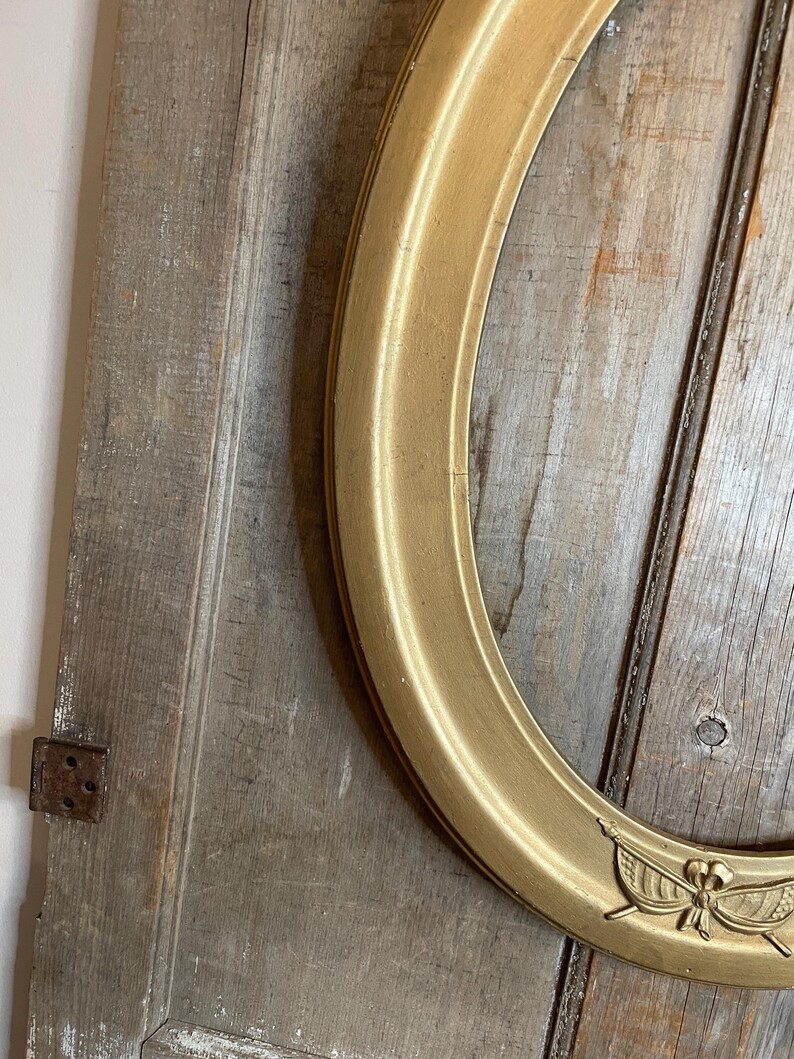 May include: A gold-colored, oval picture frame with a decorative emblem at the bottom. The frame is set against a weathered, wooden background. The frame's design is classic and elegant, suitable for displaying portraits or artwork.