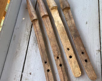 May include: Four elongated, light brown wooden tools with tapered, pointed ends and flat, rectangular handles. Each handle features two circular holes drilled through the wood. The surfaces show natural grain, texture, and signs of wear and age.