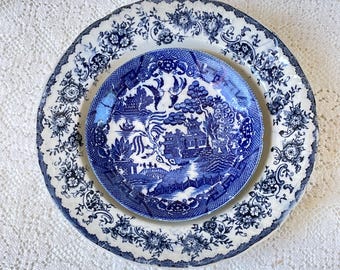 May include: Two antique ceramic plates. A larger plate with a white background and blue floral and leaf pattern surrounds a smaller plate with a blue and white landscape design. The plates are on a white lace surface.