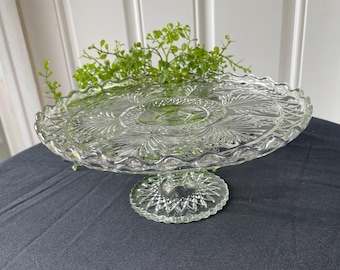 May include: A clear glass cake stand featuring a circular plate with a scalloped edge and intricate pressed patterns including fan-like motifs and a central medallion. The pedestal base has a starburst design with a textured diamond pattern.