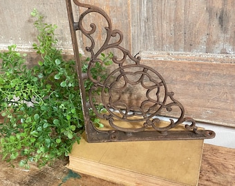 May include: A brown cast iron shelf bracket featuring an intricate scrollwork design with a circular center element. The bracket has a vertical back plate and a horizontal base plate, each with a small circular hole for mounting purposes.