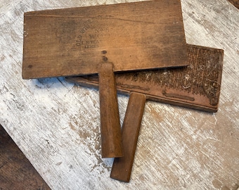 May include: Two antique wooden wool carders with wooden handles. The carders are rectangular with a dark brown finish and visible text. The handles are a lighter brown. The carders are resting on a distressed white and brown wooden surface.