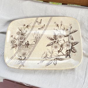 May include: A rectangular ceramic serving platter with a cream-colored base and a brown transferware pattern. The design features floral arrangements, two butterflies, and a small sailboat on water, all framed by diagonal geometric border lines.