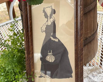 May include: Framed silhouette art of a woman in a period dress. The black silhouette is set against a cream-colored background, framed in a dark brown wooden frame. The woman is wearing a bonnet and holding a purse.