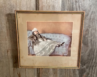 May include: Framed print of a woman in a long dress reclining on a blue sofa. The artwork is in a gold-colored frame and is set against a neutral background. The woman is wearing a hat and dark shoes.