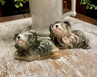 May include: Two ceramic dog figurines in a reclining position, featuring long, flowing coats with shades of dark gray, brown, and cream. Each dog has a small pink tongue visible, a black nose, and dark eyes, capturing a detailed, textured appearance.