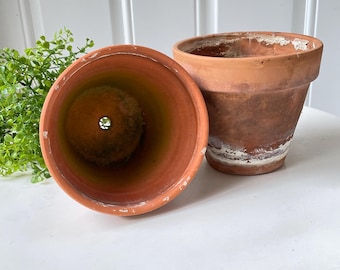 May include: Two terracotta plant pots with a weathered, earthy finish. One pot is positioned upright, showing its exterior, while the other is tilted to reveal the hollow interior and a drainage hole at the base. Both pots feature visible white mineral deposits.