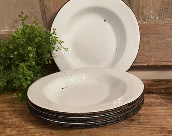 May include: A stack of white enamelware plates with black rims. The plates have a shallow, bowl-like shape. One plate is separate, and the plates are on a wooden surface. A green plant is in the background.