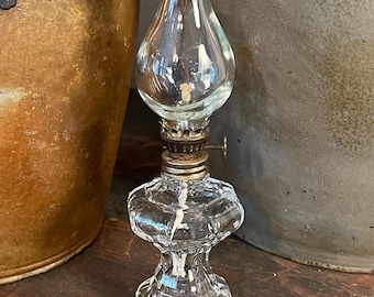 May include: A clear glass oil lamp featuring a faceted, hexagonal base and a matching glass chimney. The lamp includes a metallic burner assembly with a small adjustment knob and a visible white wick extending into the reservoir.
