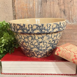 May include: A vintage stoneware mixing bowl with a cream-colored base and a blue spatter pattern. The bowl is sitting on a red book with a vintage cookbook.