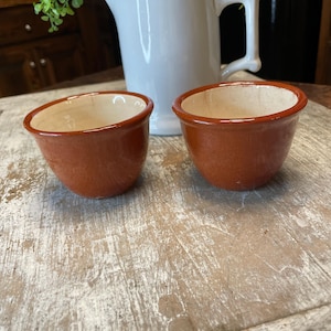 May include: Two small, terracotta-colored ceramic bowls with a light beige interior sit on a distressed, white-washed wooden surface. A white pitcher with a handle and a green plant are in the background, suggesting a rustic or farmhouse aesthetic.