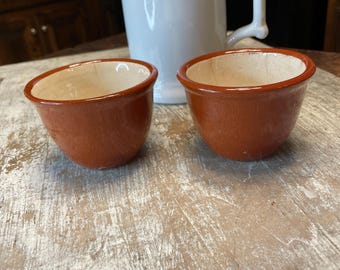 May include: Two small, terracotta-colored ceramic bowls with a light beige interior sit on a distressed, white-washed wooden surface. A white pitcher with a handle and a green plant are in the background, suggesting a rustic or farmhouse aesthetic.