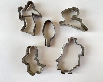 May include: Five metal cookie cutters in the shapes of people. The cutters are silver and have a worn look.