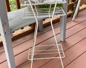 May include: A white metal plant stand featuring three tiered shelves with a distressed finish. The top shelf has a decorative perforated pattern, while the middle and bottom shelves consist of simple wire slats. The frame is constructed from thin metal rods.