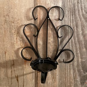May include: Black metal wall sconce with a decorative, stylized design. The sconce features a circular candle holder and ornate, curved accents. The sconce is mounted on a wooden surface.