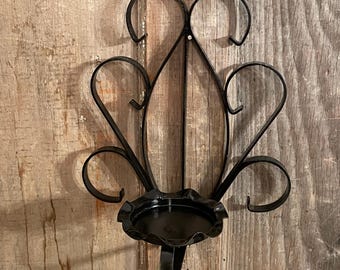 May include: Black metal wall sconce with a decorative, stylized design. The sconce features a circular candle holder and ornate, curved accents. The sconce is mounted on a wooden surface.