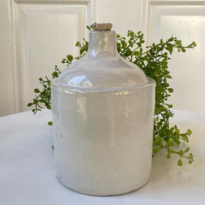May include: A cylindrical off-white ceramic jug with a smooth, glossy finish and a narrow neck topped with a small, textured cork stopper. The jug features a subtle speckled pattern throughout its surface and a slightly flared rim at the top.