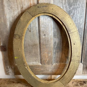 May include: Oval-shaped, antique gold-painted wooden picture frame. The frame has a distressed finish, revealing the natural wood grain beneath the gold paint. The frame is made of several pieces of wood joined together.