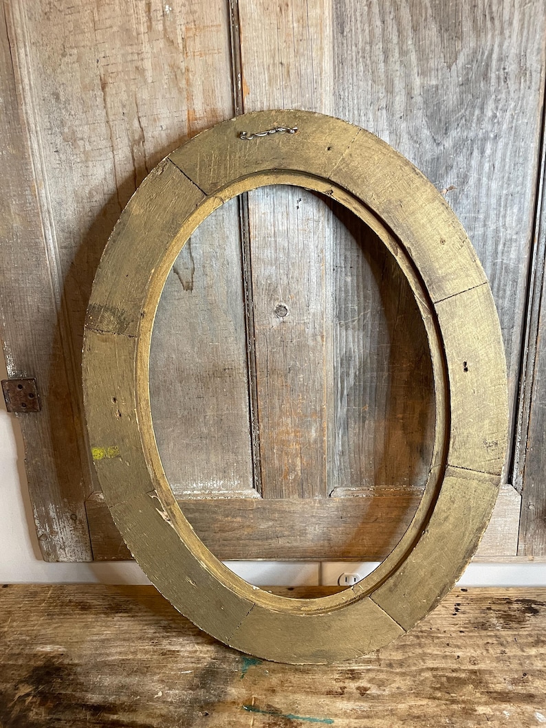 May include: Oval-shaped, antique gold-painted wooden picture frame. The frame has a distressed finish, revealing the natural wood grain beneath the gold paint. The frame is made of several pieces of wood joined together.
