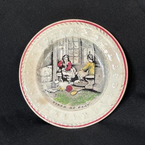 May include: A round ceramic plate with a cream-colored rim featuring a red border and embossed lettering. The center depicts a black and white illustration of two children in a room with a red ball and toy, labeled with the text TIRED OF PLAY.