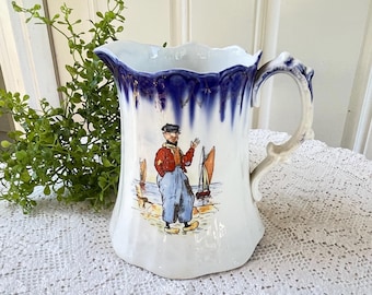 May include: A white ceramic pitcher featuring a scalloped rim with a deep blue drip glaze and gold accents. The front displays a colorful illustration of a person in a red jacket and blue trousers standing on a shore with sailboats in the background.