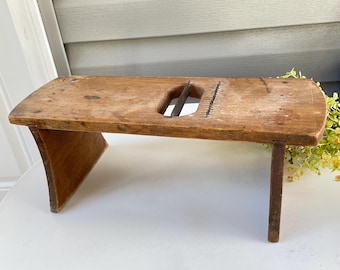 May include: A vintage wooden vegetable slicer with a rectangular top and a blade in the center. The slicer has a curved leg on one side and a straight leg on the other. The wood has a natural, aged appearance.