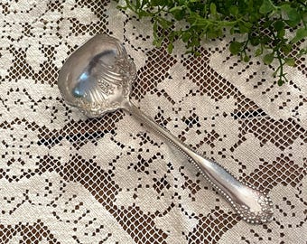 May include: A silver-toned metal serving spoon featuring a wide, shell-shaped bowl with intricate embossed floral and scrollwork detailing. The handle is slender with a beaded edge and a decorative floral motif at the base of the stem.