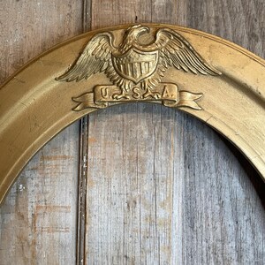May include: A gold-colored, oval-shaped frame with an embossed eagle and shield design at the top. The banner below the eagle reads "USA". The frame is set against a weathered, wooden background.